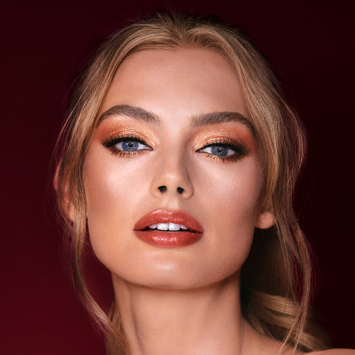 A light-tone model with blue eyes wearing shimmery copper and gold eye makeup with black eyeliner, glowy bronzed cheeks, and orange-red lipstick with gloss on top. 