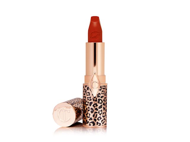 An open orange-red matte lipstick in a golden, cheetah-print, metallic packaging with its lid next to it. 