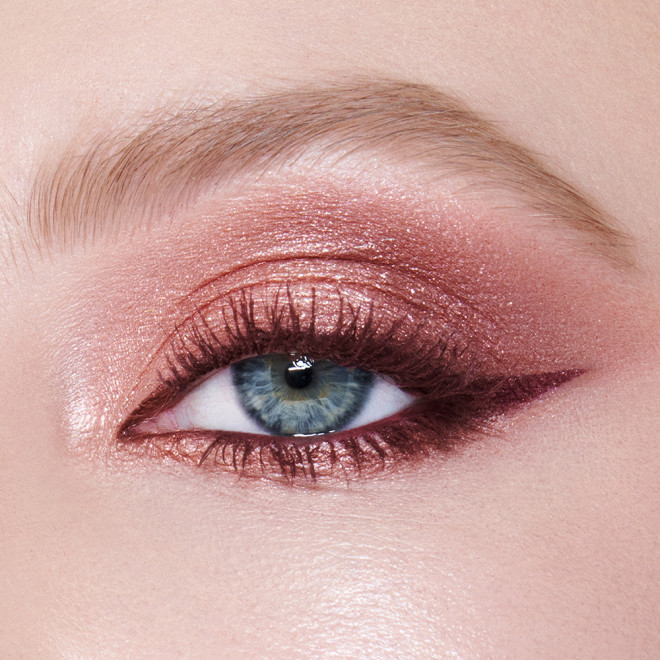 Einzelne Nahaufnahme des Auges eines Models mit hellem Hautton und blauen Augen, das ein Augen-Make-up in Perlmutt-Roségold, Altrosa, Beerenbraun und Rosenknospenrosa mit beerenbraunem Eyeliner und dunkelbrauner Mascara trägt. 