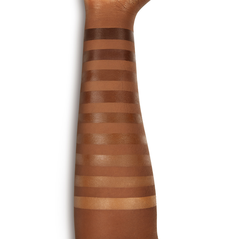 Deep-tone model's arm with swatches of nine cream concealers in shades of dark beige, light brown, dark brown, and black-brown. 