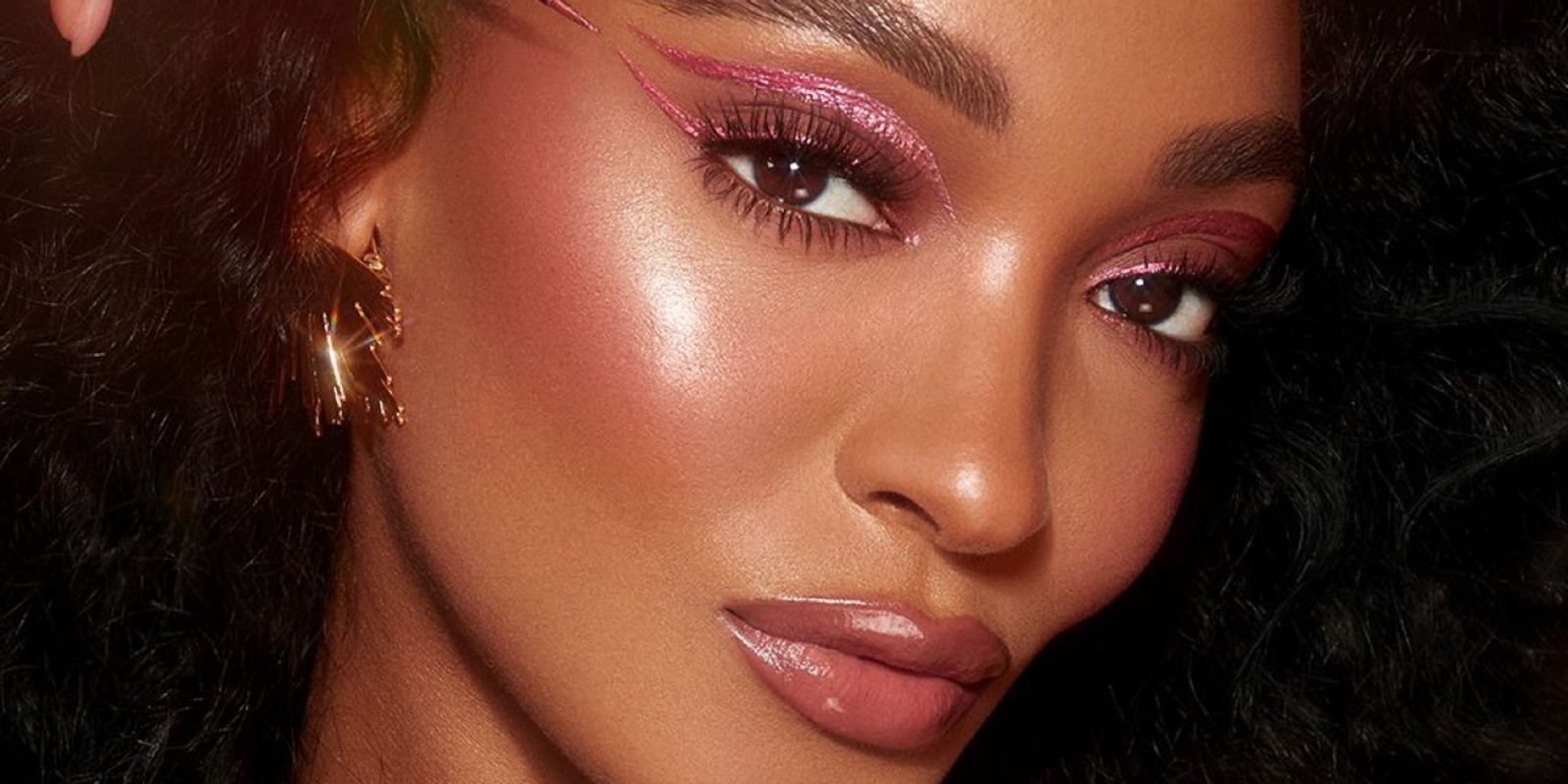 A deep-tone model with brown eyes applying a glittery vivid, rose pink eyeliner while wearing full-glam dewy and glowy pink makeup.