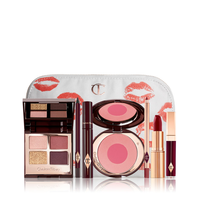 A makeup bag with 7 makeup products, an open, mirrored-lid eyeshadow palette in matte and shimmery gold and red shades, an open black eyeliner pencil, a mascara in a dark-crimson colour scheme, a red lipstick with a matching lip liner pencil, vampy-red lip gloss, and an open two-tone blush in cool pink. 