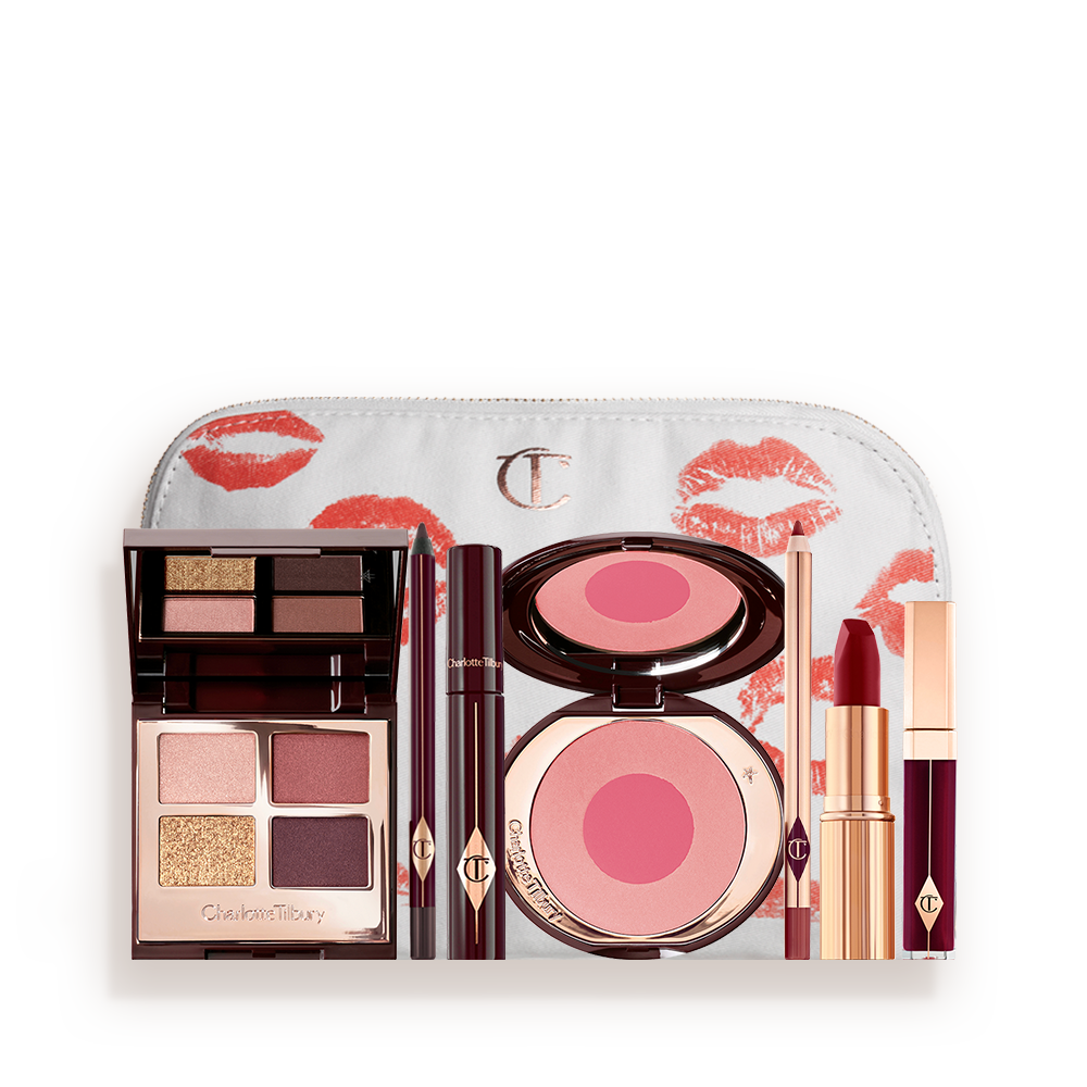 A makeup bag with 7 makeup products, an open, mirrored-lid eyeshadow palette in matte and shimmery gold and red shades, an open black eyeliner pencil, a mascara in a dark-crimson colour scheme, a red lipstick with a matching lip liner pencil, vampy-red lip gloss, and an open two-tone blush in cool pink. 