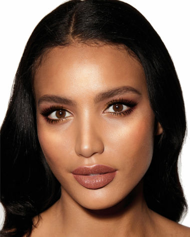 A deep-tone model with brown eyes wearing shimmery copper and gold eye makeup with black eyeliner, glowy bronzed cheeks, and pinkish-brown lipstick with gloss on top. 