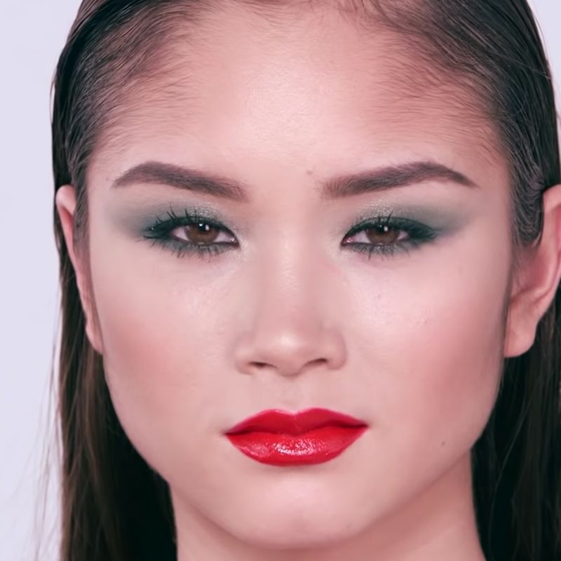 A light-tone model with brown eyes wearing smokey grey and teal eyes makeup with a soft pink blush and glossy vibrant red lips.
