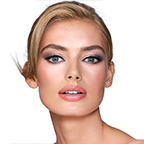 A fair-tone model with blue eyes wearing shimmery lilac and champagne eye makeup with nude pink blush and coral lipstick with gloss on top.