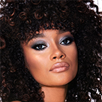 A deep-tone brunette model with brown eyes wearing smokey silvery eyeshadow with bronzed cheeks and glossy nude-peach lips. 