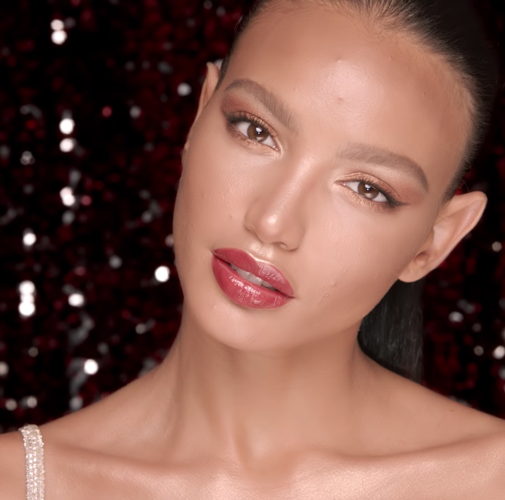 A medium-tone model with a glowy face base with shimmery rose gold and brown eye makeup with glossy berry-red lips. 