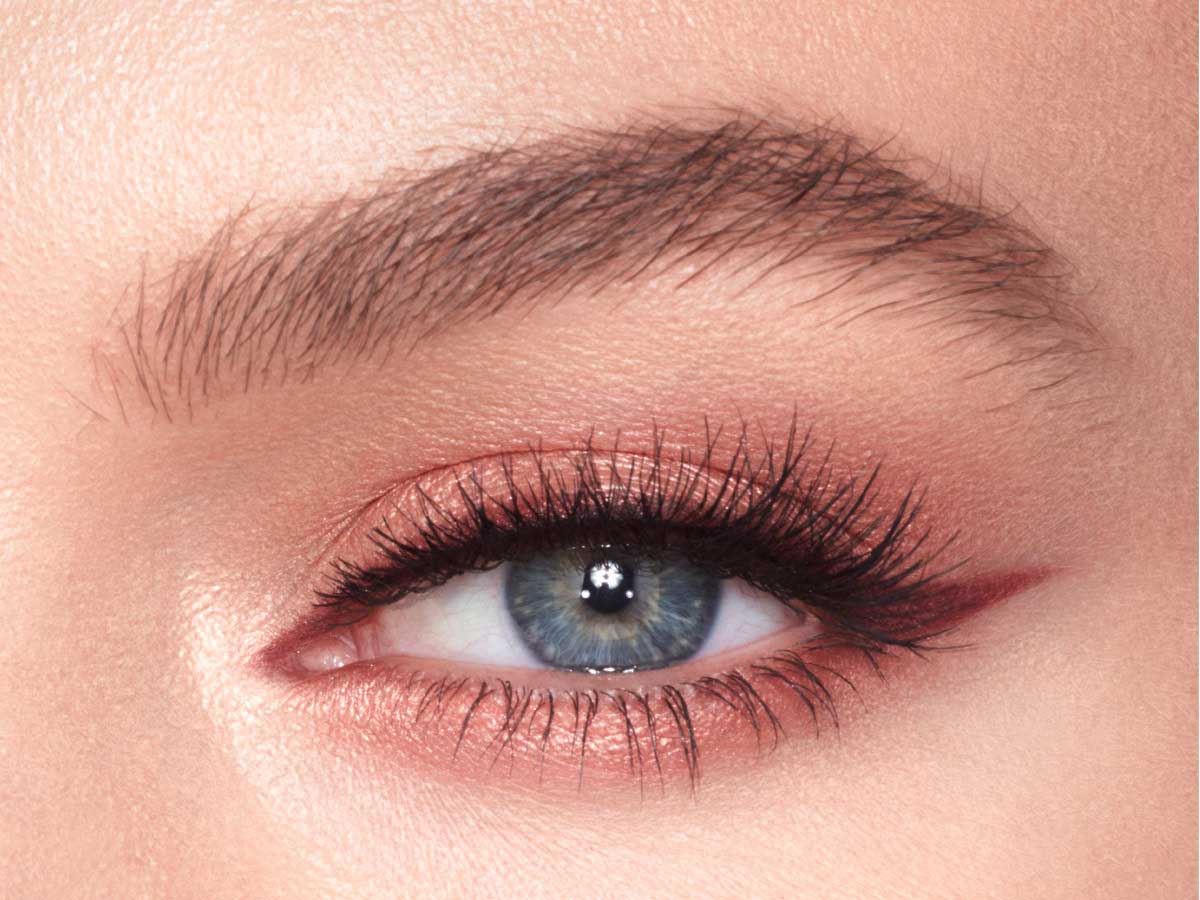 Eye close-up of a model with blue eyes wearing shimmery peach and rose gold eye makeup with soft, berry-brown eyeliner. 