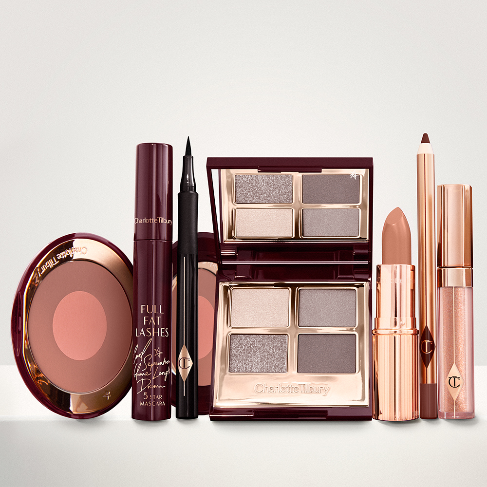 An open two-tone blush in cool-toned brown and rose gold with a mascara, eyeliner pen, quad eyeshadow palette with shimmery and matte grey, beige and silver shades, an open lipstick in nude red, lip liner pencil in taupe-brown, and a lip gloss in nude pink. 