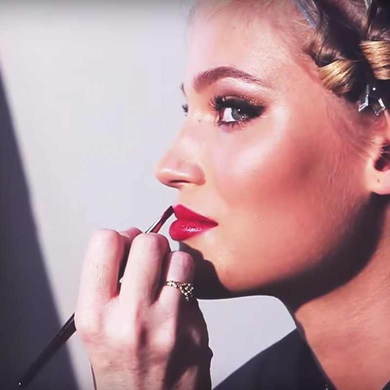 A light-tone blonde model with blue eyes wearing full-glam makeup, getting a blue-toned red lipstick applied.