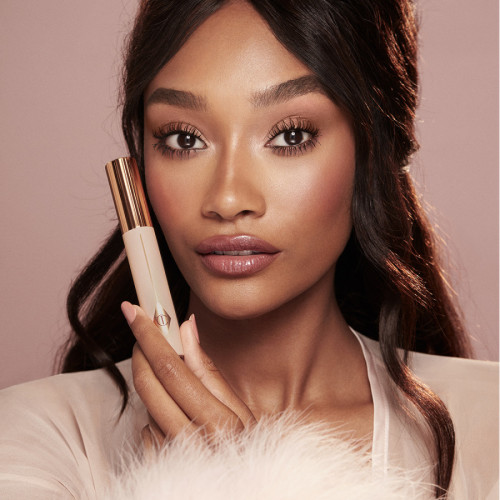 A deep-tone model with brown eyes wearing nude pink lipstick with muted pink blush, and lengthening mascara in a berry-brown shade while holding the mascara bottle beside her face.