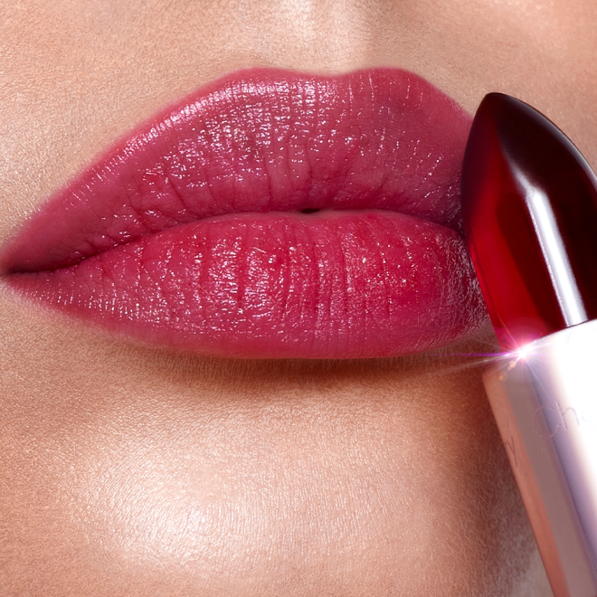 Lips close-up of a fair-tone model wearing a subtle nude red lipstick while holding a glowy, crystal-like red lipstick against her lips. 