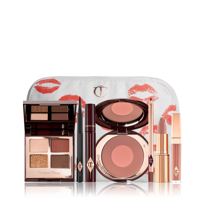 A white makeup pouch with an open two-tone blush in cool-toned brown and warm pink with a mascara, eyeliner pen, quad eyeshadow palette with shimmery and matte brown and golden shades, an open lipstick in nude peach, lip liner pencil in taupe-brown, and a lip gloss in nude pink. 