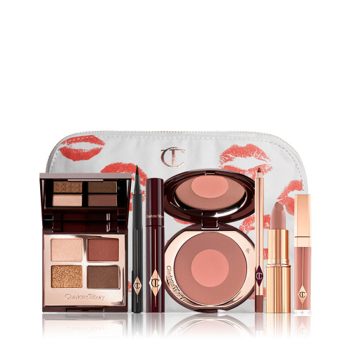 A white makeup pouch with an open two-tone blush in cool-toned brown and warm pink with a mascara, eyeliner pen, quad eyeshadow palette with shimmery and matte brown and golden shades, an open lipstick in nude peach, lip liner pencil in taupe-brown, and a lip gloss in nude pink. 