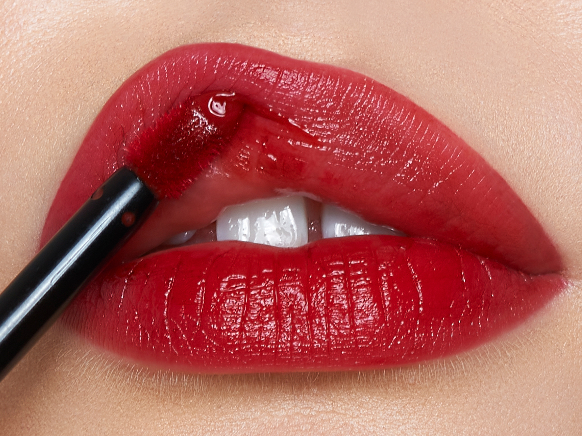 Lips close-up of a light-tone model applying a vibrant, poppy-red lip tint with a doe-foot brush applicator. 