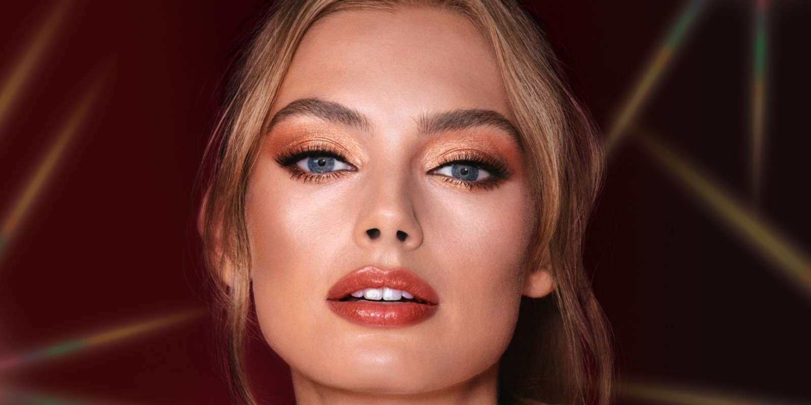 A fair-tone model with blue eyes wearing shimmery copper and gold eye makeup with black eyeliner, glowy bronzed cheeks, and pinkish-brown lipstick with gloss on top. 