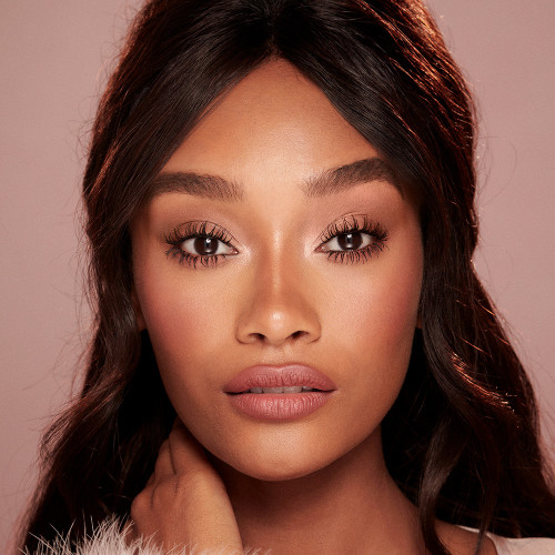 A deep-tone model with brown eyes wearing nude pink lipstick with muted pink blush, and lengthening mascara in a berry-brown shade.