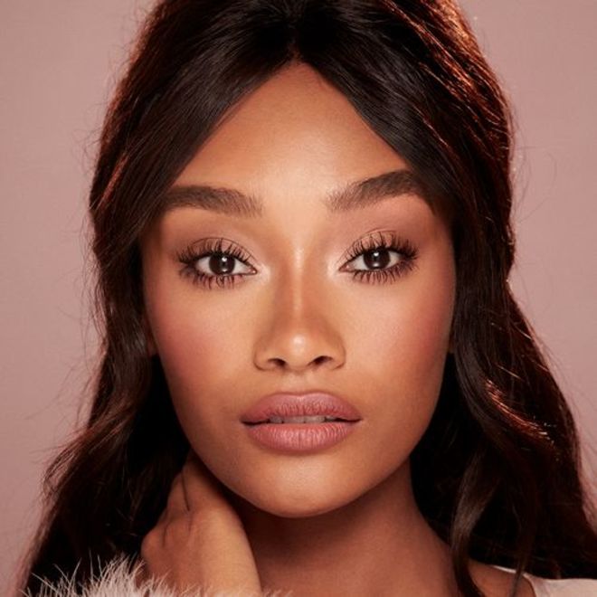 Deep-tone model with brown eyes wearing nude pink matte lipstick with warm pink blush, and volumizing and lengthening mascara in black colour.