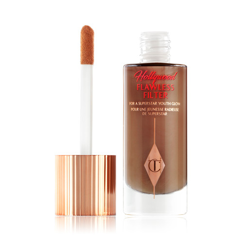 An open, luminous primer in a dark brown shade in a glass bottle with its gold and white doe-foot applicator next to it.
