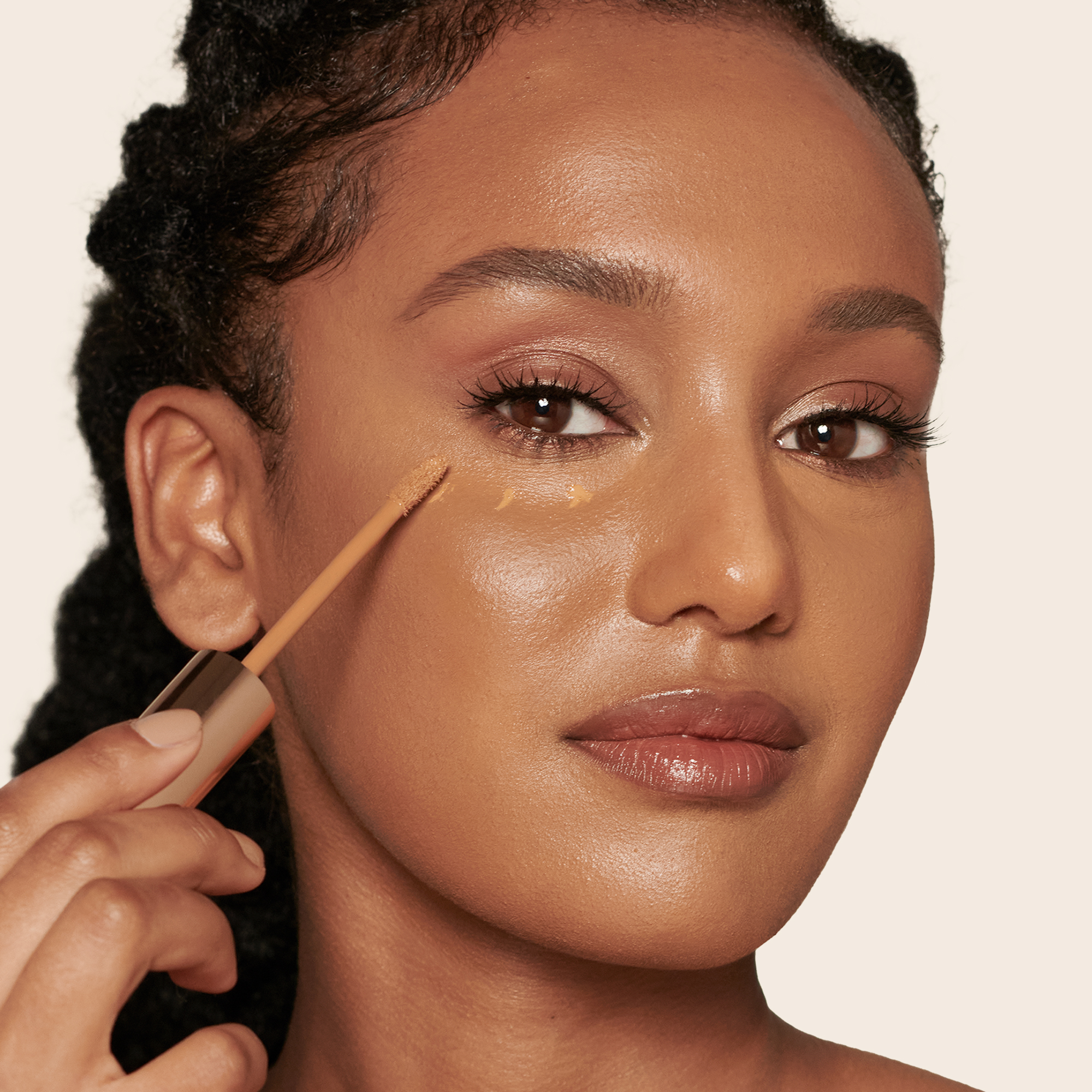 Deep-tone model wearing dewy, soft-glam makeup applying glowy, concealer in a dark brown shade. 