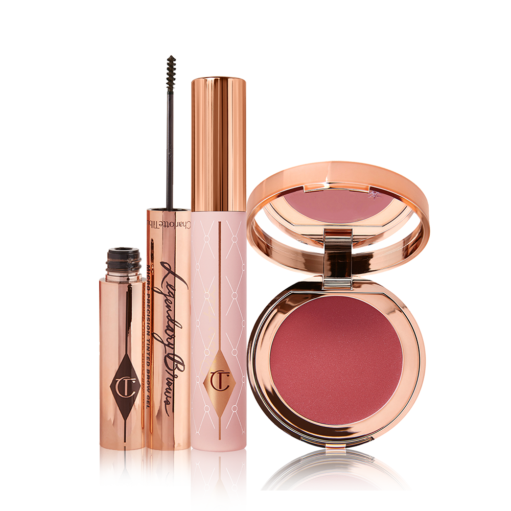 An eyebrow tint with its thin applicator for precise application next to it, mascara in a nude pink-coloured tube with a gold-coloured lid, and a lip and cheek tint compact with a mirrored-lid in a berry-rose shade.