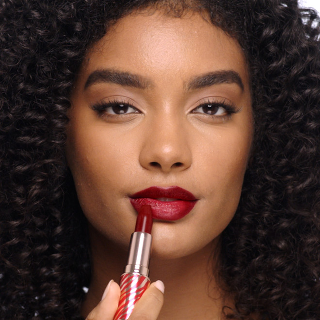 Deep-tone model with brown eyes applying a rich berry-rose lipstick with lips lined with a lip liner in a matching shade. 