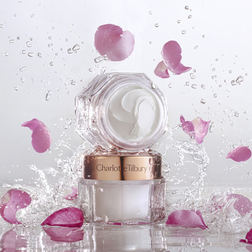 Two pearly-white face creams, one with a gold-coloured lid and the other without, in glass jars with sparkling water and rose petals falling on them. 
