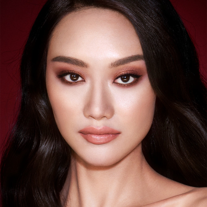 A fair-tone model with brown eyes wearing smokey brown and gold eye makeup with warm pink blush and glossy nude-pink lips.