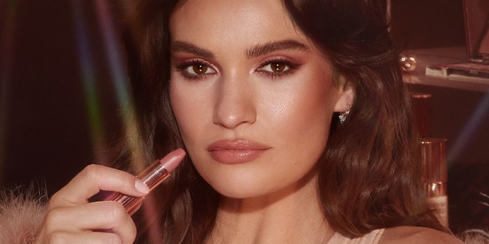 Lily James wearing dewy, full-glam makeup in nude pink while wearing nude pink matte lipstick while holding the lipstick.