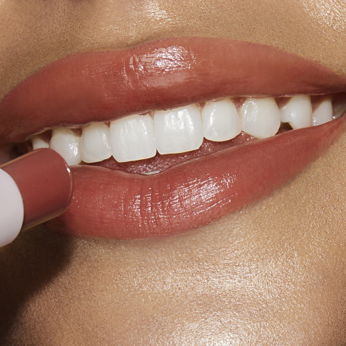 Lips close-up of a medium-tone model applying a moisturising lipstick balm in a peachy nude shade with a high-shine finish.