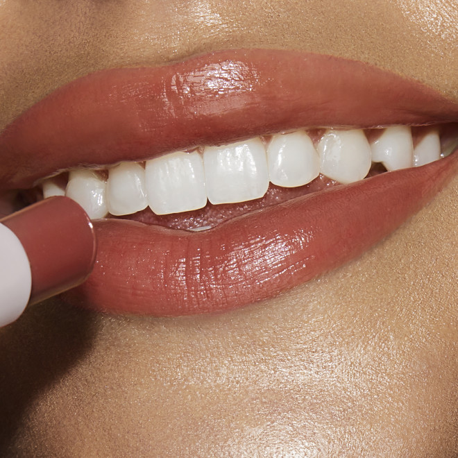 Lips close-up of a medium-tone model applying a moisturising lipstick balm in a peachy nude shade with a high-shine finish.