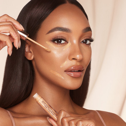 Deep-tone model with brown eyes applying a radiant concealer on the high points of her face for a lifted look and wearing nude lip gloss and subtle eye makeup.