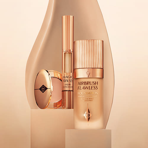 An open powder compact in a light-medium shade, a concealer, and a foundation in a frosted glass bottle, all in golden-coloured packaging. 