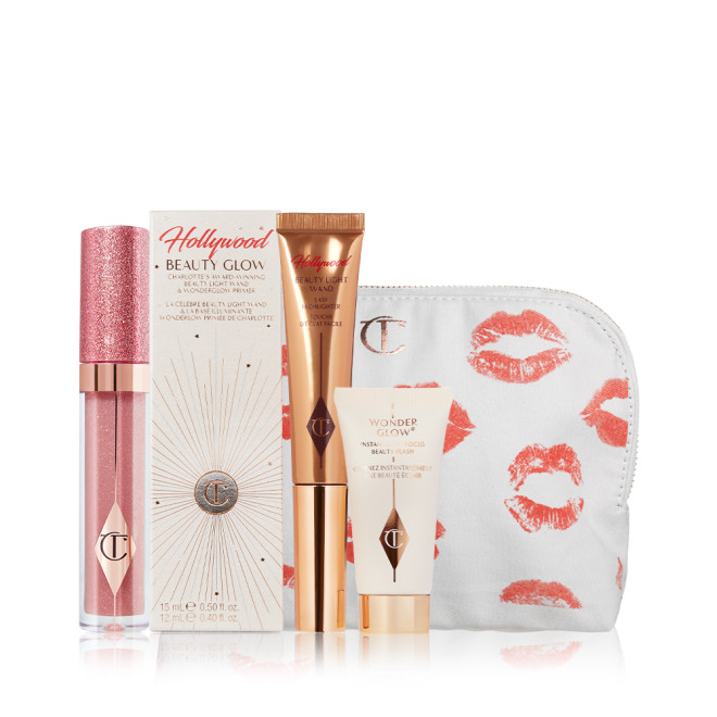 Nude pink shimmery lip gloss in a glass tube, glowy primer in a white-coloured box, liquid highlighter wand in a bronze-gold-coloured tube, and a white-coloured makeup pouch with kiss prints all over. 
