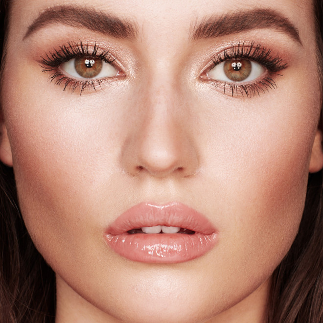A light-tone, brunette model with hazel eyes wearing shimmery, rose gold makeup with nude pink lip gloss. 