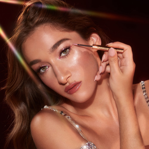A light-tone model with brown eyes applying a glittery vivid, rose pink eyeliner while wearing full-glam dewy and glowy pink makeup.