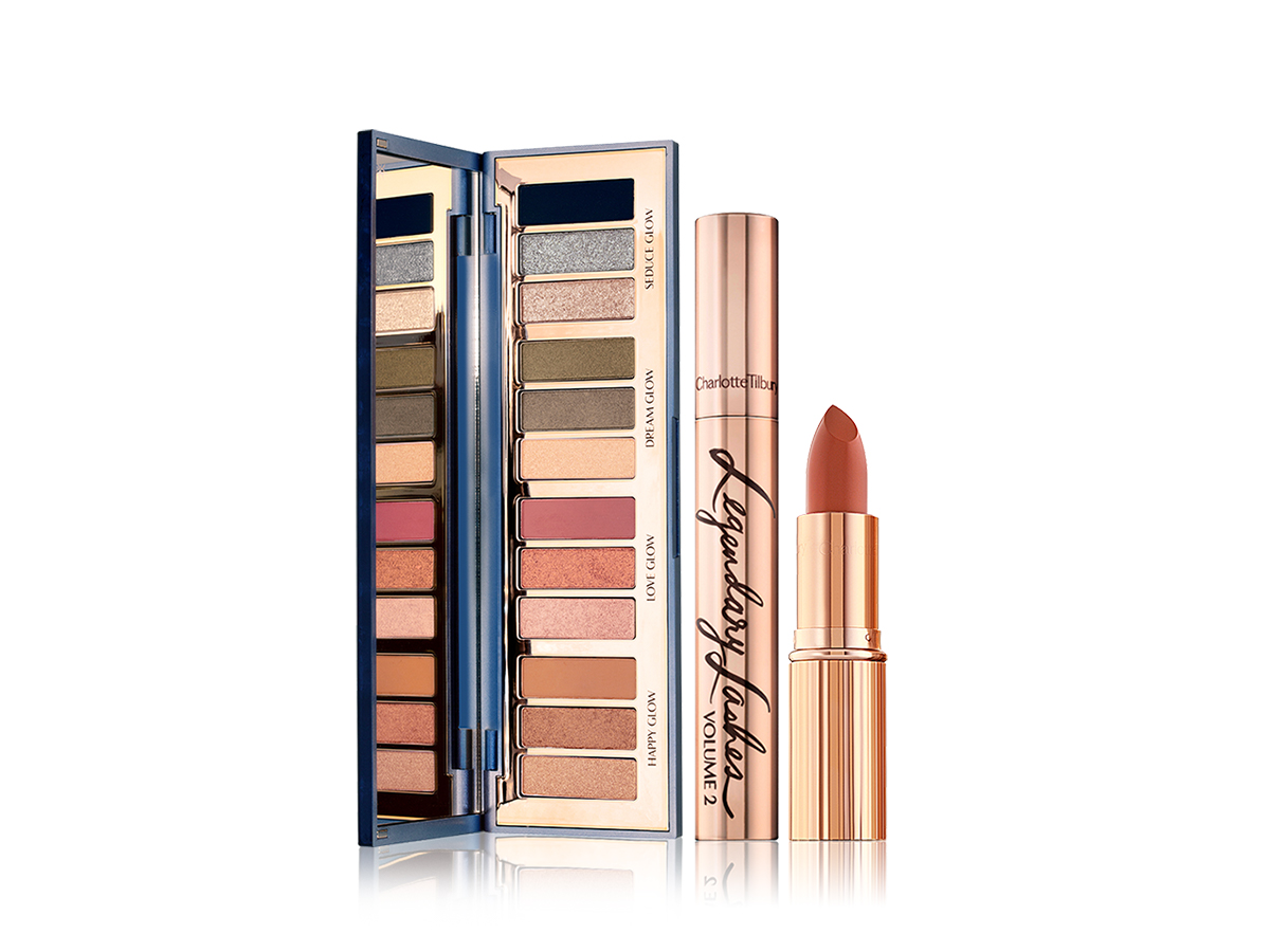 An open, mirrored-lid eyeshadow palette with eyeshadows in shades of pink, brown, peach, golden, blue, and green, along with a mascara with gold packaging, and an open lipstick in a medium-dark coral shade.