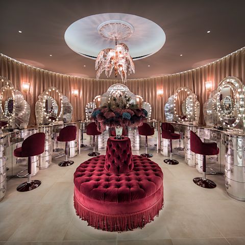Un magasin de maquillage aux thèmes rouge et doré avec des chaises tournantes et des tables de maquillage miroir avec des lumières à LED. 