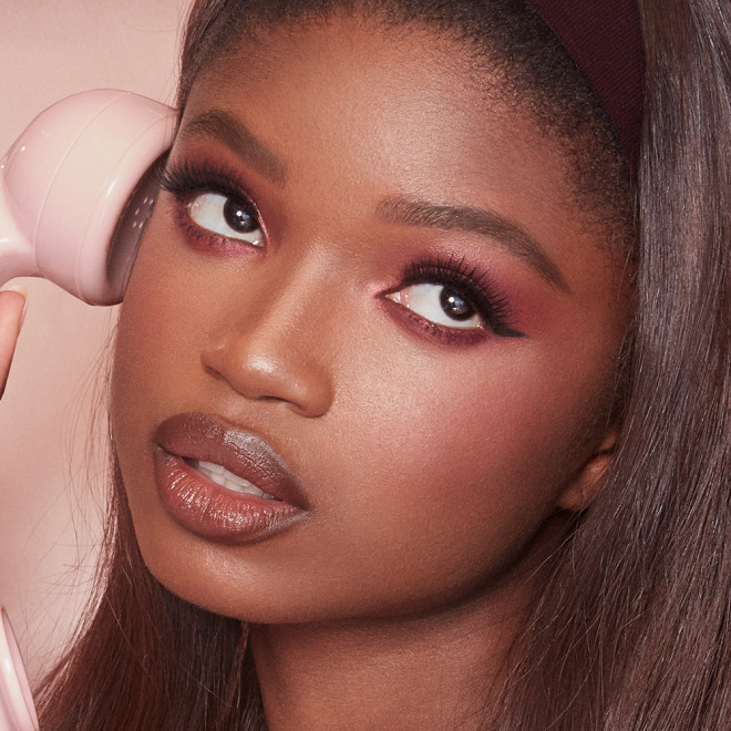 Close-up of a brunette, deep skin model, wearing soft-glam, berry-pink makeup, and holding a nude-pink phone. 
