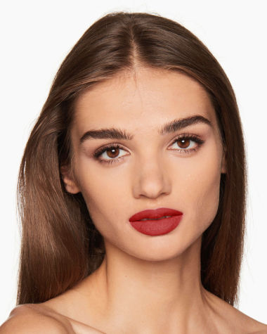 A medium-tone model with brown eyes wearing a muted, matte brick red lipstick. 