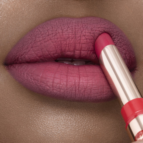 Lips close-up of a deep-tone model applying a soft pink-red lipstick with a matte finish.