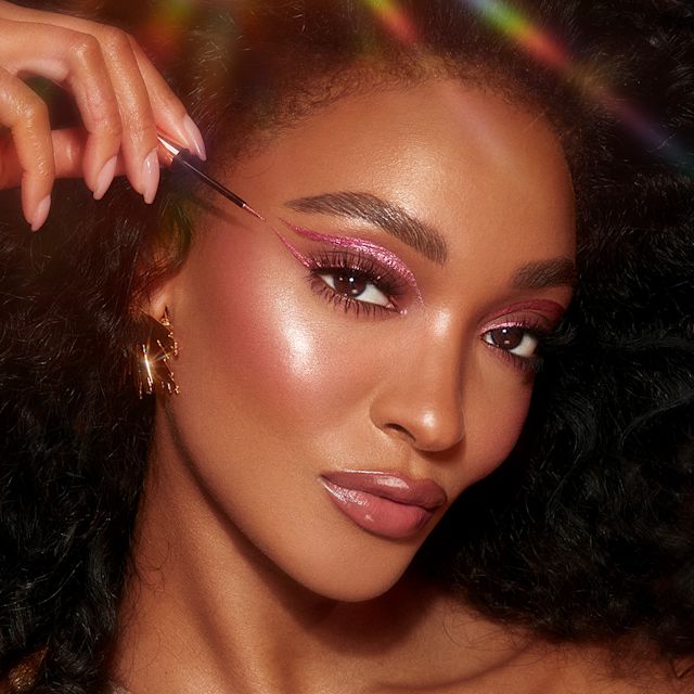 A deep-tone model with brown eyes applying a glittery vivid, rose pink eyeliner while wearing full-glam dewy and glowy pink makeup.