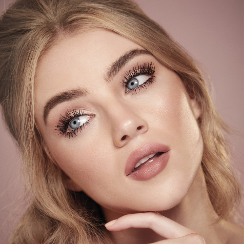 A fair-tone model with blue eyes wearing nude pink lipstick with muted pink blush, and lengthening mascara in a berry-brown shade. 