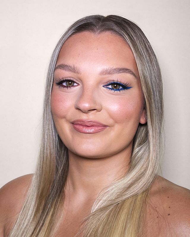 Close-up of a fair-tone model with green eyes wearing soft pinkish-peach matte lipstick with brownish-pink blush, and brown and beige eye makeup with blue eyeliner on her lower waterline.