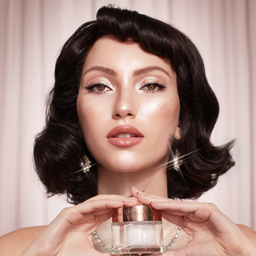 Fair-tone brunette model wearing full-glam, glowy nude peach makeup while holding a jar of pearly-white face cream with a gold-colorued lid.