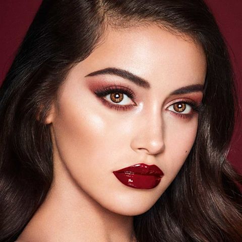 A fair-tone model with brown eyes wearing reddish-plum eye makeup, glowy face base, and a vampy-red lipstick with a satin-finish. 