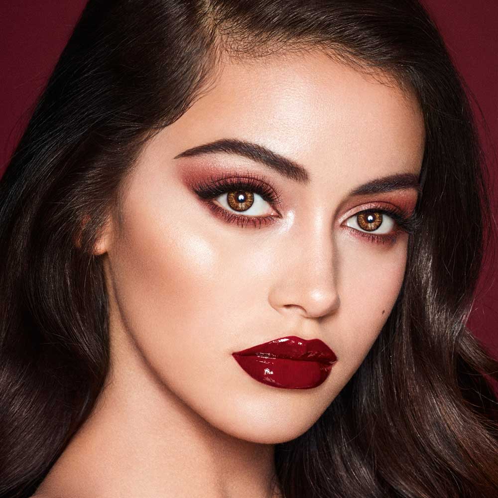 A fair-tone model with brown eyes wearing reddish-plum eye makeup, glowy face base, and a vampy-red lipstick with a satin-finish. 