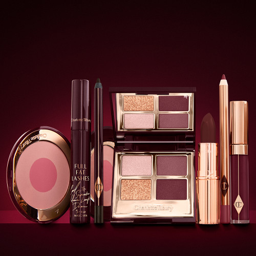 An open two-tone blush in fuchsia and warm pink with a mascara, eyeliner pencil, quad eyeshadow palette with shimmery and matte brown and golden shades, an open lipstick in crimson red, lip liner pencil in wine, and a lip gloss in vampy red. 