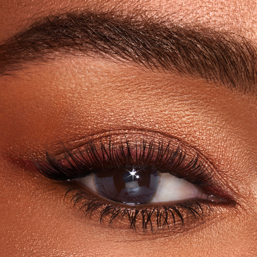 Single-eye close-up of a deep-tone model with brown eyes wearing shimmery topaz and brown eyeshadow with dark brown eyeliner pencil. 